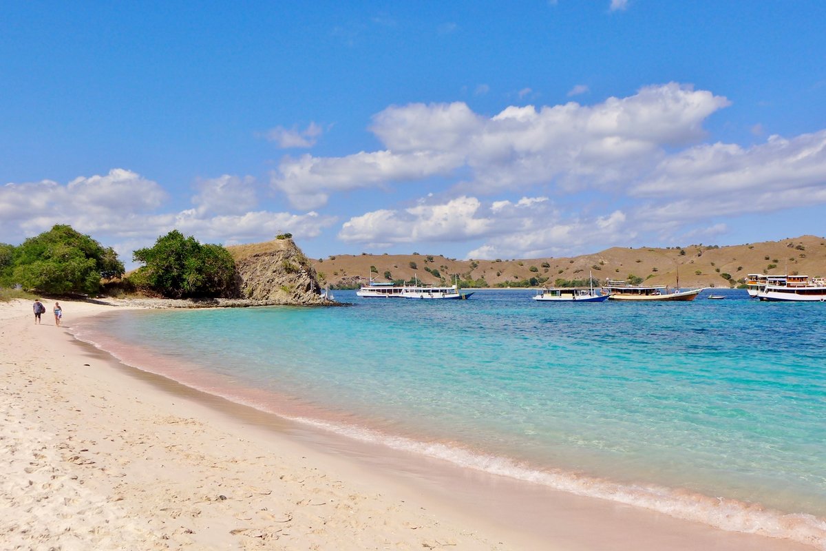 Pink sand and turquoise water at Pink Beach in Komodo, with boats anchored offshore and dry hills behind the bay