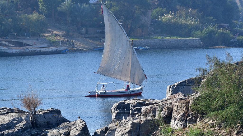 A felucca with its sail up carrying passengers on the Nile at Aswan, with rocky banks in the foreground.
