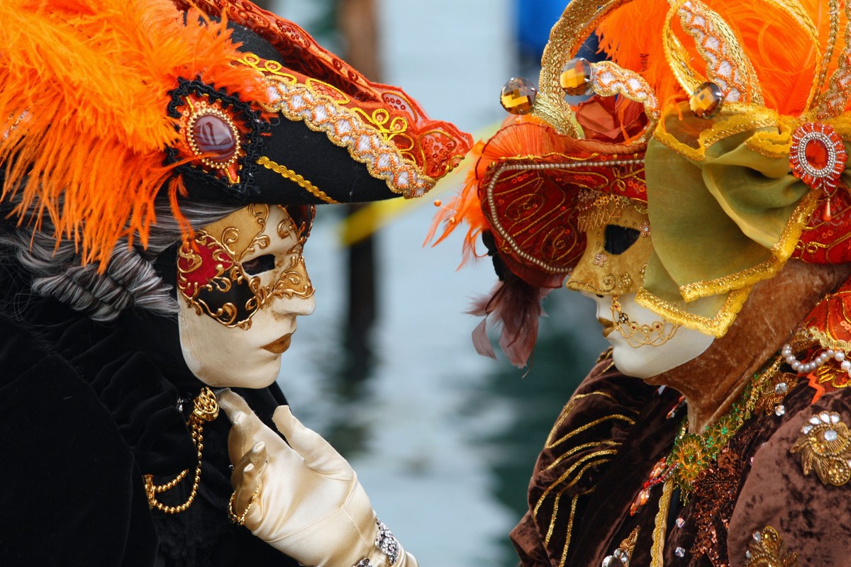 wo Venice Carnival performers in ornate gold and orange masks and feathered costumes facing each other