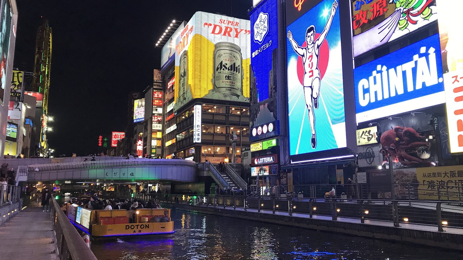 The Glico running man sign and neon billboards over the canal at Dotonbori, Osaka, at night, with Ebisubashi bridge and a tour boat