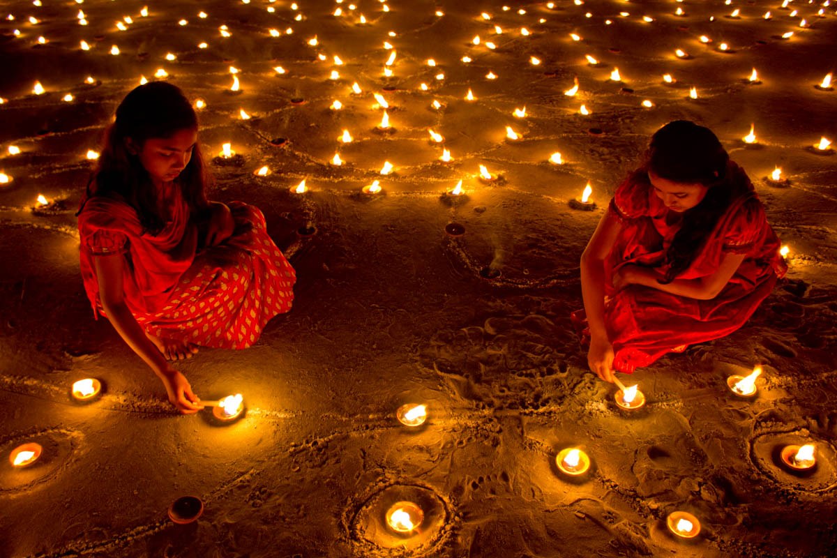 Two girls in red dresses lighting small oil lamps on the ground during Diwali, surrounded by hundreds of flames