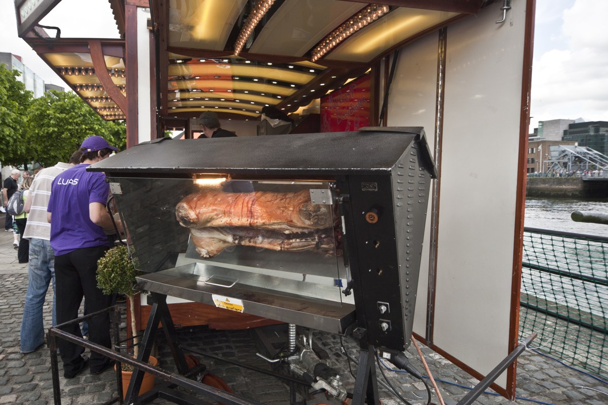 A whole hog roasting on a spit at an outdoor food festival stall beside a river