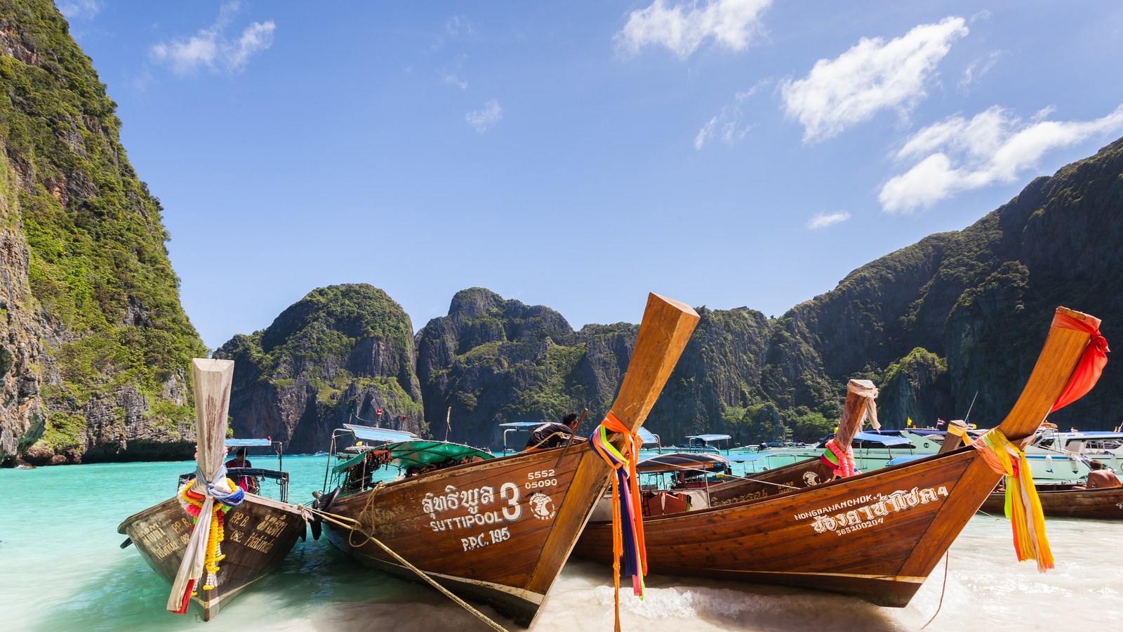 Wooden longtail boats with ribbon-tied prows in the turquoise shallows of Maya Bay, Ko Phi Phi Lee, Thailand, below jungle-covered limestone karsts