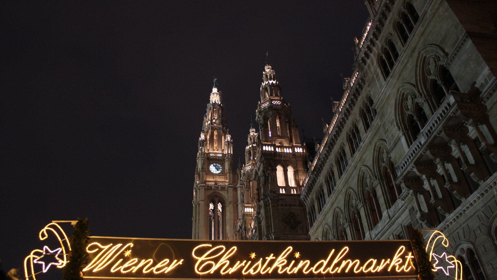 The illuminated Wiener Christkindlmarkt entrance sign with the Rathaus towers behind it at night in Vienna
